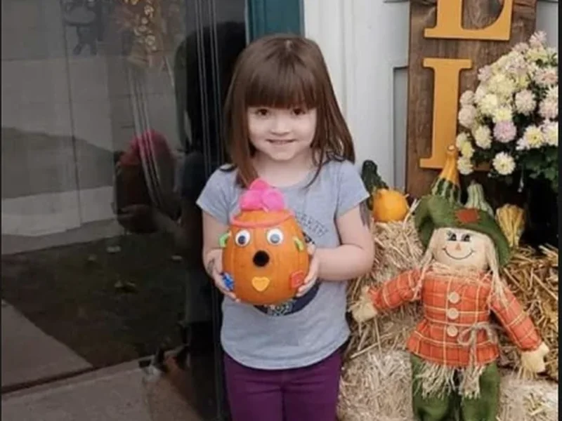 A school-age girl on the front step holding a decorated pumpkin, beside the FALL sign and a scarecrow.
