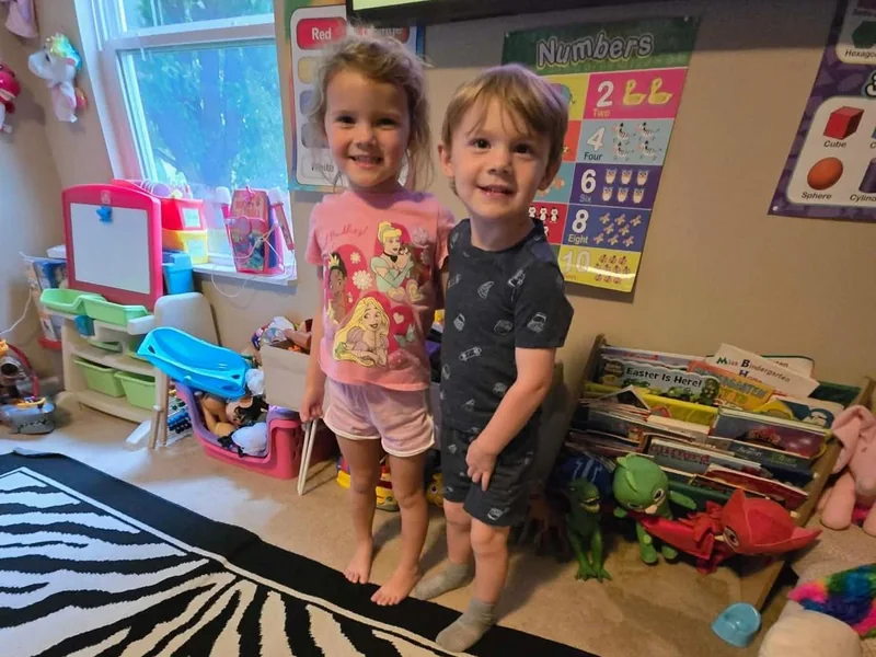 Two toddlers standing on the zebra rug in the playroom, in front of the bookshelf and the numbers poster.
