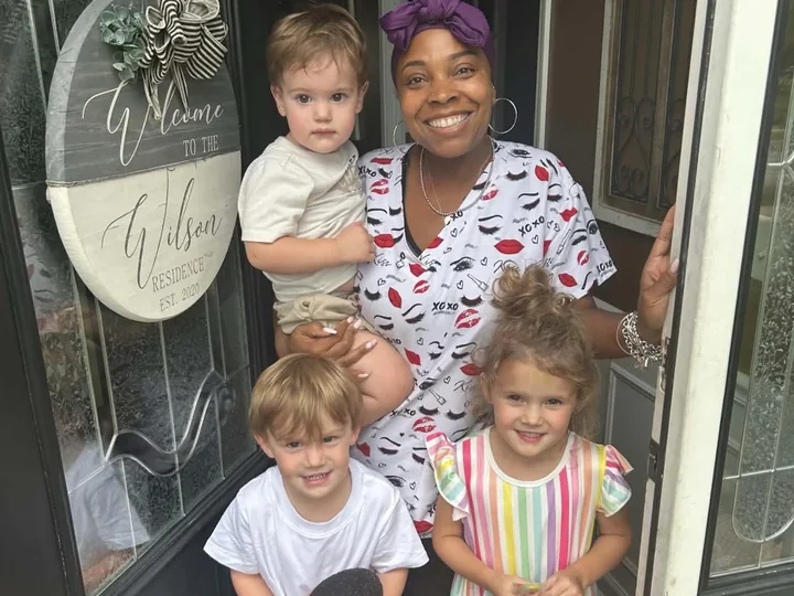 Colia Wilson at her front door holding a toddler, with two more children in front of her, beside the Wilson Residence welcome sign.