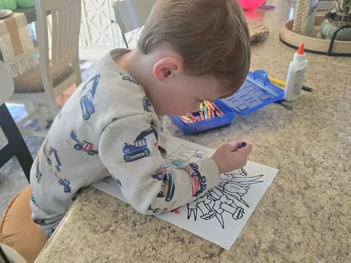 A boy coloring a dinosaur page at the dining table, with a box of crayons and a bottle of glue beside him.