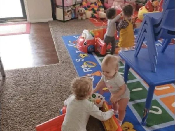Three toddlers playing on the alphabet rug in the playroom with a red ride-on car.