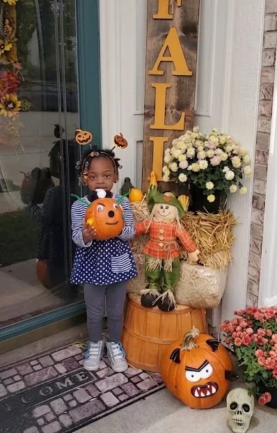 A girl on the front step holding a jack-o'-lantern craft, beside a wooden FALL sign, a scarecrow, mums, and a carved pumpkin.
